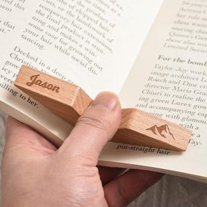 May include: A wooden bookmark with the name "Jason" engraved on it. The bookmark also features a mountain design. It is placed between the pages of a book.