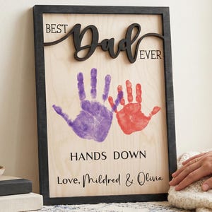 Puede incluir: Cartel enmarcado en madera con las palabras "BEST Dad EVER" en escritura negra. El cartel presenta huellas de manos moradas y rojas y el texto "HANDS DOWN Love, Mildred & Olivia".