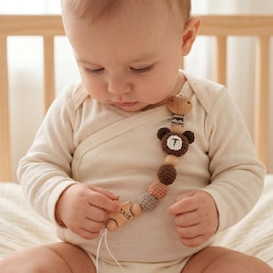 Op de afbeelding: Een baby in een lichtbeige romper houdt een houten fopspeenclip vast. De clip heeft een bruin berenontwerp met de letter "T", houten kralen en een stervormig houten stuk met de tekst "axis". Op de achtergrond is een houten bed te zien.