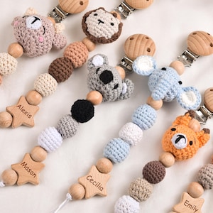 Op de afbeelding: Een verzameling handgemaakte fopspeenclips voor baby's. Elke clip heeft een gehaakt dierenhoofd, houten kralen en een houten ster met een gegraveerde naam. De dieren zijn onder meer een beer, leeuw, koala, olifant en vos.