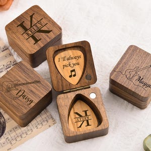 Puede incluir: Cajas de madera para púas de guitarra con grabados personalizados. Una caja está abierta, revelando una púa con "I'll always pick you" y una nota musical. Otras cajas presentan nombres y contornos de guitarra. Las cajas son cuadradas y de madera oscura.