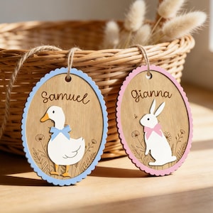 Könnte beinhalten: Zwei hölzerne Osterei-förmige Anhänger mit den Namen "Samuel" und "Gianna". Der Samuel-Anhänger hat einen blauen Rand und eine Entenillustration. Der Gianna-Anhänger hat einen rosa Rand und eine Hasenillustration.