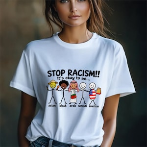 May include: A white t-shirt featuring a graphic of five stick figures holding hands. The text reads STOP RACISM!! It's okay to be... followed by labels for each figure: asian, black, arab, normal, and american, with corresponding cultural icons.