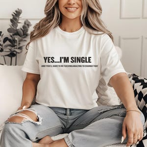 May include: White t-shirt with the text "YES... I'M SINGLE" in black, with a second line of text below. The shirt is worn with blue jeans. The overall theme is casual wear.