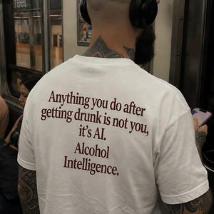 Peut inclure: Un t-shirt blanc à manches courtes avec un texte marron foncé imprimé au dos : Anything you do after getting drunk is not you, it's AI. Alcohol Intelligence. Le vêtement est porté par une personne ayant des tatouages visibles sur les bras.