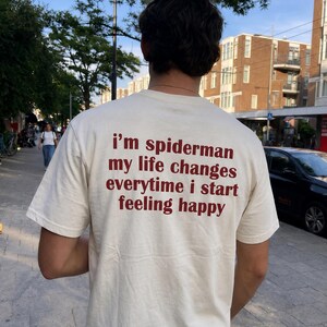Op de afbeelding: Een crèmekleurig T-shirt met korte mouwen en donkerrode tekst op de achterkant: i'm spiderman my life changes everytime i start feeling happy. Het shirt heeft een ronde hals en een standaard pasvorm, waardoor de tekst duidelijk leesbaar is.