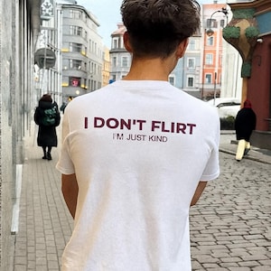Może przedstawiać: Biała koszulka z napisem "I DON'T FLIRT I'M JUST KIND" w bordowym kolorze. Koszulka jest noszona przez osobę idącą po brukowanej ulicy. W tle widać budynki i inne osoby.