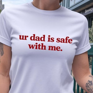 May include: White t-shirt with the red text "ur dad is safe with me." The shirt has short sleeves and a rolled cuff. The text is centered on the chest.