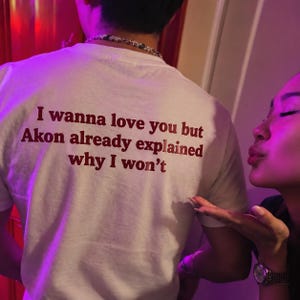 Op de afbeelding: Een wit t-shirt met donkerrode tekst op de achterkant die luidt: I wanna love you but Akon already explained why I won't. Het shirt heeft een ronde hals en wordt gedragen met een zilveren ketting die zichtbaar is in de nek.