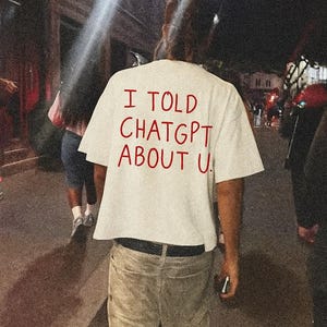 May include: A white t-shirt with the red text "I TOLD CHATGPT ABOUT U." The shirt is worn by a person walking down a street at night. The person is wearing khaki shorts and white socks.