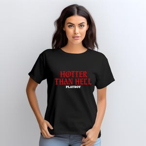 Puede incluir: Camiseta negra con el texto rojo "HOTTER THAN HELL" y la palabra "PLAYBOY" en blanco. La camiseta es de cuello redondo y tiene mangas cortas. La modelo lleva la camiseta.