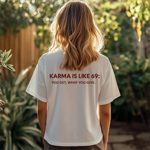 May include: A white short-sleeved t-shirt featuring dark red text on the back that reads: KARMA IS LIKE 69: YOU GET, WHAT YOU GIVE. The shirt has a crew neckline and a relaxed fit, displaying the printed message clearly across the upper back area.