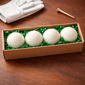 May include: Four white golf balls in a brown cardboard box filled with green shredded paper. A white golf glove and a wooden tee are also visible on a dark wood surface. The golf balls have a dimpled surface.