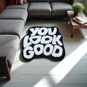 以下が含まれることがあります： 太字の「YOU LOOK GOOD」という文字が入った白黒のラグ。白い木の床に置かれ、背景には灰色のソファと小さな木製のテーブルがあります。装飾的な家のアクセントです。