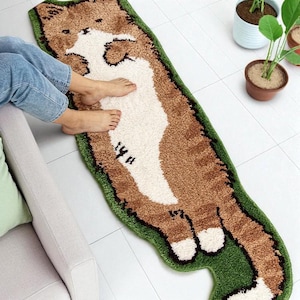 Puede incluir: Una alfombra con forma de gato en marrón, blanco y verde. La alfombra muestra un gato tumbado boca arriba, con un borde verde. Está colocada sobre un suelo de baldosas blancas. El texto "14 A" es visible. Mide unos 150 cm de largo.