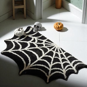 Può includere: Un tappeto nero e bianco a forma di ragnatela con un teschio e piccole zucche. Il tappeto è nero con un disegno a ragnatela bianca. Una zucca bianca, un teschio e una zucca arancione sono sul pavimento.