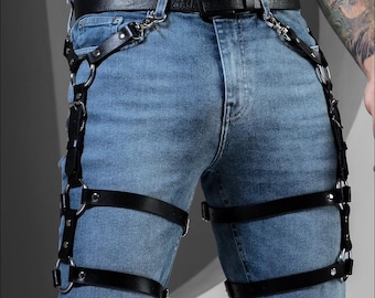 May include: Black leather harness with silver-tone metal rings and buckles. The harness is worn over blue jeans and includes straps that connect to a black belt. The design features multiple straps and rings for a unique look.