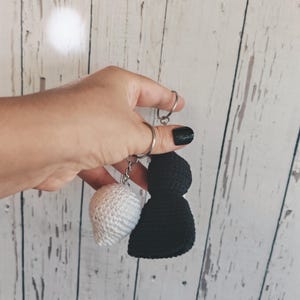May include: Two crochet keychains attached to a silver metal ring. One keychain is a small, round, white sphere made of yarn, and the other is a larger, black, bell-shaped crochet piece. Both items are connected by silver metal jump rings.