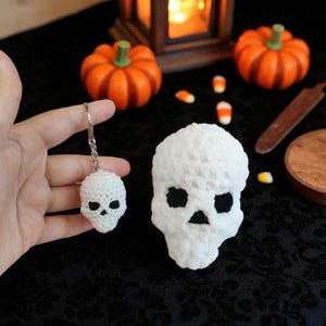 May include: A white crocheted skull keyring next to a larger white crocheted skull. The skulls have black eye and nose openings. The image has Halloween decorations, including orange pumpkins and candy corn.