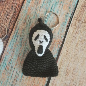 May include: A crochet keychain featuring a black hooded figure with a white mask. The mask has black embroidered eyes and an open mouth. The figure is attached to a silver metal split ring, creating a small, textured, handmade accessory.