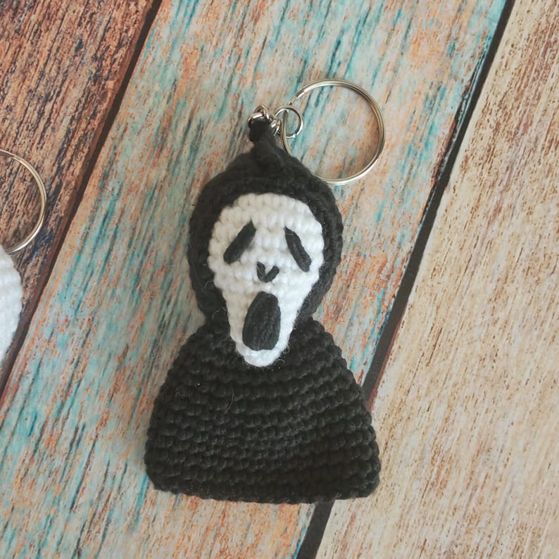May include: A crochet keychain featuring a black hooded figure with a white mask. The mask has black embroidered eyes and an open mouth. The figure is attached to a silver metal split ring, creating a small, textured, handmade accessory.