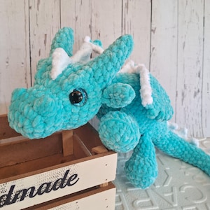 K&ouml;nnte beinhalten: Ein t&uuml;rkisfarbener geh&auml;kelter Drachen-Pl&uuml;schtier mit wei&szlig;en Akzenten an H&ouml;rnern und R&uuml;cken. Der Drache hat gro&szlig;e, funkelnde blaue Augen und liegt auf einer Holzkiste. Der Pl&uuml;sch besteht aus weichem, strukturiertem Garn.