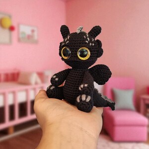 May include: A black crocheted dragon with large, golden eyes and small wings. The handmade plush toy has gray accents on its ears, feet, and tail. The dragon is sitting on a hand, with a pink background.
