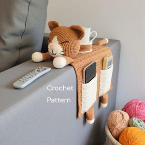 May include: Crocheted cat-shaped sofa armrest organizer in shades of brown and white. Features a sleeping cat head, pockets for remote controls and phones, and a space for a mug. The text "Crochet Pattern" is visible.