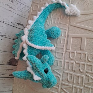 K&ouml;nnte beinhalten: Ein t&uuml;rkisfarbener geh&auml;kelter Drachen mit wei&szlig;en Akzenten. Der Drache hat einen langen Hals, Fl&uuml;gel und einen Schwanz mit einer flauschigen wei&szlig;en Spitze. Die Augen des Drachen sind schwarz, und er liegt auf einer strukturierten Oberfl&auml;che.