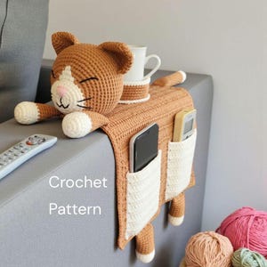 Puede incluir: Un organizador de sofá con forma de gato tejido a ganchillo en tonos marrón y blanco. El organizador presenta una cabeza de gato, patas y cola, con bolsillos para un teléfono y un mando a distancia. Una taza y un posavasos blancos están encima. El texto "Crochet Pattern" es visible.