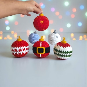 May include: A hand holding a red crocheted Christmas ornament with gold accents, surrounded by other handmade ornaments. The ornaments are red, white, blue, and green, with various designs. The background has blurred lights.