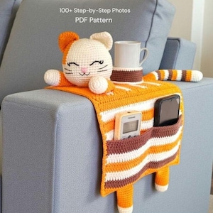 May include: Crocheted cat-shaped sofa armrest organiser in orange, white, and brown. Features a cat head, striped body, pockets for a remote and phone, and a cup holder. Text reads "100+ Step-by-Step Photos PDF Pattern."