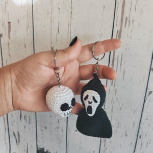 May include: Two crochet keychains featuring horror-themed designs. One is a small white skull with black stitched eye sockets and teeth. The other is a black hooded figure with a white face, black eyes, and an open mouth, both on metal rings.