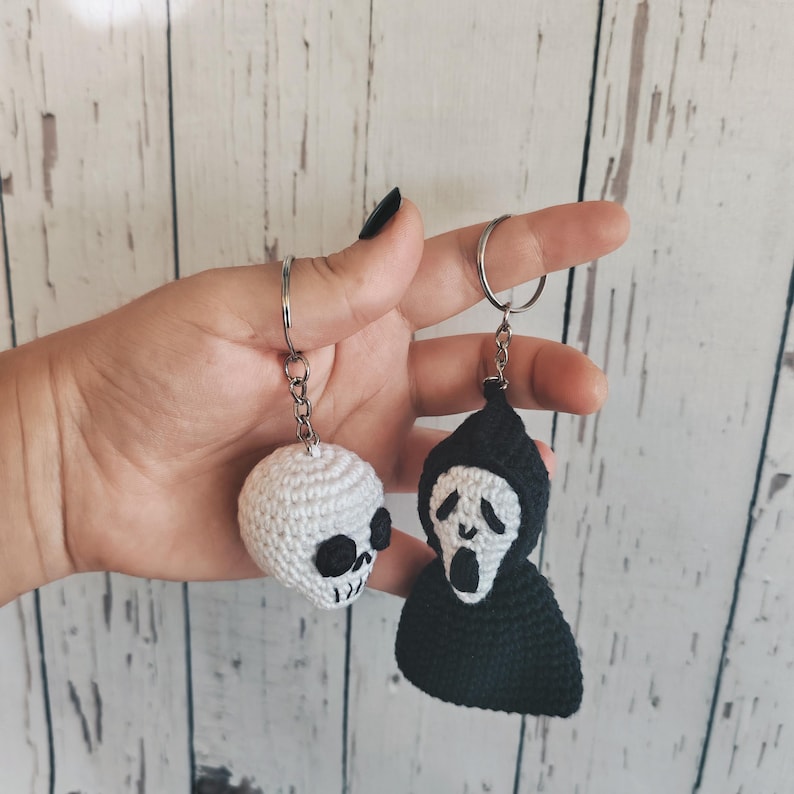 May include: Two crochet keychains featuring horror-themed designs. One is a small white skull with black stitched eye sockets and teeth. The other is a black hooded figure with a white face, black eyes, and an open mouth, both on metal rings.