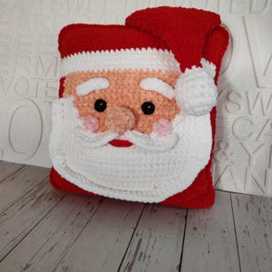 May include: A crocheted cushion in the shape of Santa Claus, featuring a red hat and trim, a white beard and moustache, a peach-coloured face, black eyes, and pink cheeks. A festive Christmas decoration.