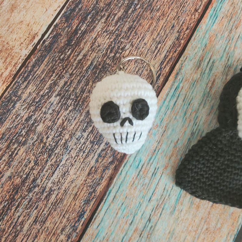 May include: A white crocheted skull keychain featuring black yarn circles for eye sockets, a small black stitched nose, and vertical black lines representing teeth. The skull is attached to a circular silver metal split ring for keys.