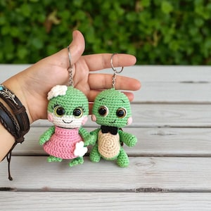 Puede incluir: Dos llaveros de tortuga hechos a mano con ganchillo. Una tortuga lleva un vestido rosa y una flor blanca, la otra tiene un caparazón beige y una pajarita negra. Ambas tienen ojos grandes y expresivos y están unidas a llaveros plateados.