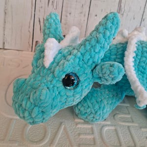K&ouml;nnte beinhalten: Ein handgefertigter t&uuml;rkisfarbener H&auml;keldrachen mit wei&szlig;en Akzenten. Der Drache hat gro&szlig;e, glitzernde schwarze Augen und ein weiches, strukturiertes Aussehen. Die Fl&uuml;gel und H&ouml;rner sind wei&szlig; und kontrastieren mit dem t&uuml;rkisfarbenen K&ouml;rper. Dieses Pl&uuml;schtier ist eine skurrile Kreation.