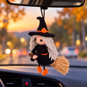 Puede incluir: Un adorno de bruja de ganchillo colgado de un espejo retrovisor. La figura tiene un sombrero puntiagudo negro con un detalle de calabaza naranja, largo cabello de lana gris, un vestido negro con detalle naranja en la cintura, zapatos naranjas y una escoba de lana beige.