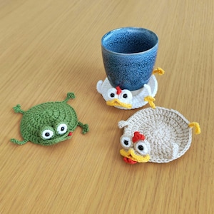 Könnte beinhalten: Drei handgefertigte Häkeluntersetzer in Form von Zeichentricktieren. Einer ist grün und froschartig, die anderen beiden sind huhnförmig, einer weiß und einer beige. Eine blaue Keramiktasse steht auf einem der Hühneruntersetzer.
