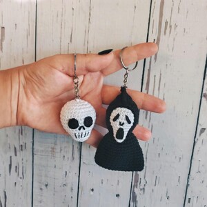 May include: Two crochet keychains featuring horror-themed designs. One is a white skull with black embroidered eyes and mouth. The other is a black hooded figure with a white face, black eyes, and a black mouth, both attached to silver metal rings.