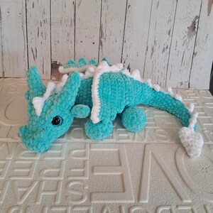 K&ouml;nnte beinhalten: Ein t&uuml;rkisfarbener H&auml;keldrache mit wei&szlig;en Akzenten. Das handgefertigte Pl&uuml;schtier hat ein detailliertes Design mit langem Schwanz, kleinen Beinen und einem Kopf mit H&ouml;rnern. Der Drache ist aus weichem, strukturiertem Garn gefertigt und hat schwarze Augen.