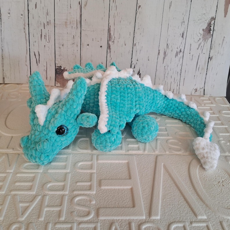 K&ouml;nnte beinhalten: Ein t&uuml;rkisfarbener H&auml;keldrache mit wei&szlig;en Akzenten. Das handgefertigte Pl&uuml;schtier hat ein detailliertes Design mit langem Schwanz, kleinen Beinen und einem Kopf mit H&ouml;rnern. Der Drache ist aus weichem, strukturiertem Garn gefertigt und hat schwarze Augen.