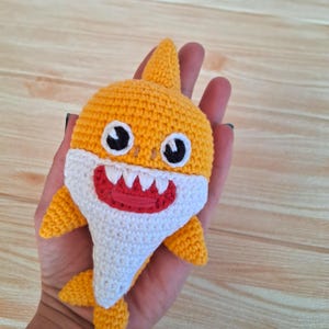 May include: A handmade crocheted shark toy in yellow, white, and red. The shark features black eyes, a white belly, and a red mouth with white teeth. The toy has a textured appearance.