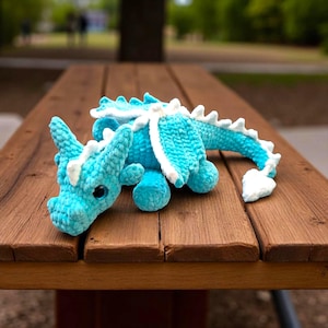 May include: A turquoise crochet dragon toy with white accents, including wings and spikes. The plush dragon is lying on a wooden table, showcasing its detailed craftsmanship and soft texture. The dragon's eyes are black.