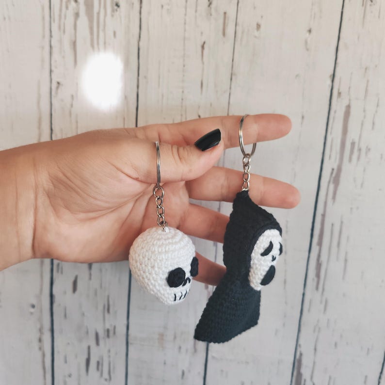 May include: Two crochet keychains featuring a white skull with black eye sockets and a black-hooded figure with a white face, black eyes, and an open mouth. Each item is attached to a silver-toned metal split ring with a short chain link connector.