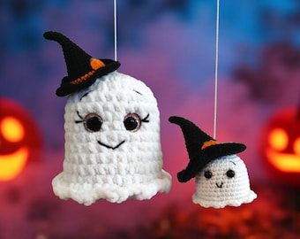 May include: Two white crochet ghosts with black witch hats, orange accents, and smiling faces hang against a blurred Halloween background. One ghost is larger than the other. The ghosts are suspended by white strings.