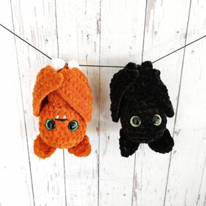 Pode incluir: Dois morcegos de pelúcia pendurados em uma corda. Um é laranja com detalhes brancos e o outro é preto. Ambos têm olhos verdes iridescentes grandes e são feitos de um material texturizado, perfeitos para o Halloween.
