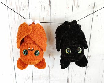 Puede incluir: Dos murciélagos de peluche colgando de una cuerda. Uno es naranja con detalles blancos y el otro es negro. Ambos tienen grandes ojos verdes iridiscentes y están hechos de un material texturizado, ideal para Halloween.