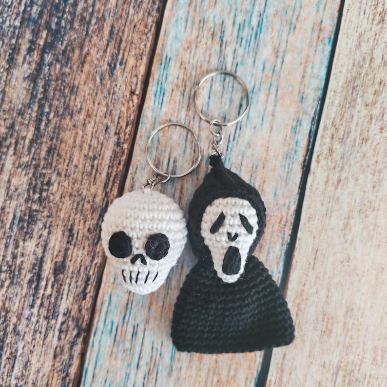May include: Two crochet keychains featuring a white skull with black embroidered eyes and teeth, and a figure wearing a black hooded cloak with a white face mask that has black eye, nose, and mouth details, both attached to silver metal rings.
